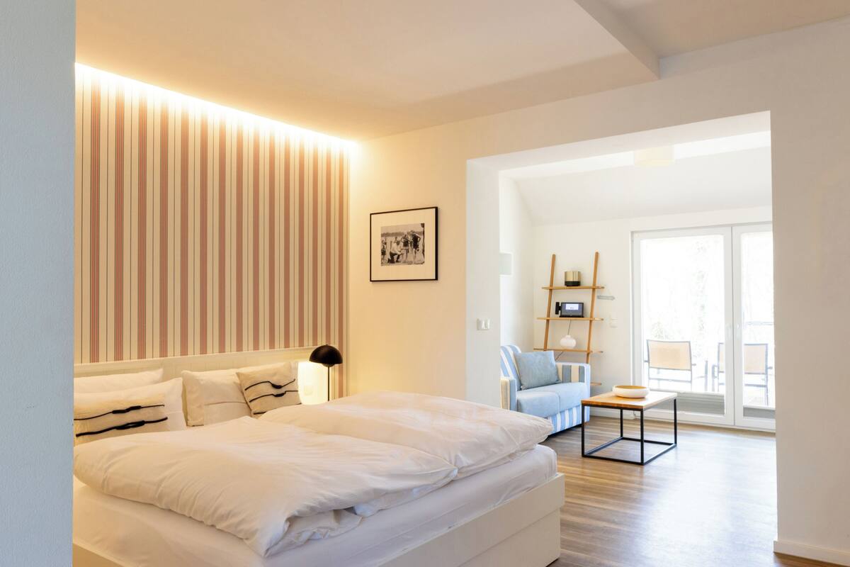 A modern hotel room with a large bed, a bedside table, a lamp, a framed picture on the wall, and a view of the outdoor area.