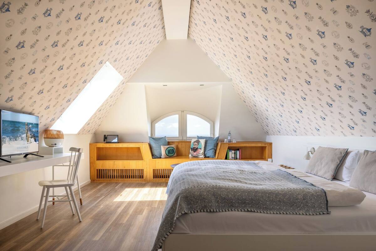 A bedroom with a sloped ceiling, a bed, a desk, a chair, and a window with a view.