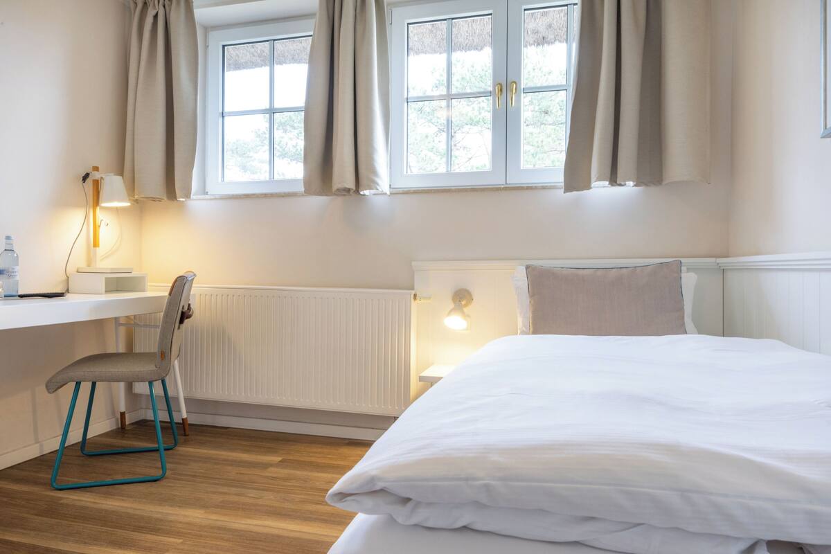 A bedroom with a bed, a desk with a chair, a radiator, and a window with curtains.