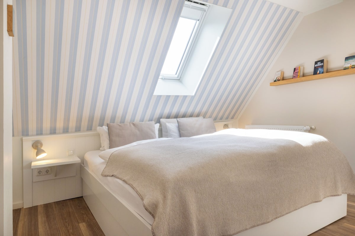 A bedroom with a sloped ceiling, a skylight, a bedside table, a bed with a beige blanket, and a shelf with books.
