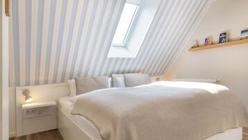 A bedroom with a sloped ceiling, a skylight, a bedside table, a bed with a beige blanket, and a shelf with books.