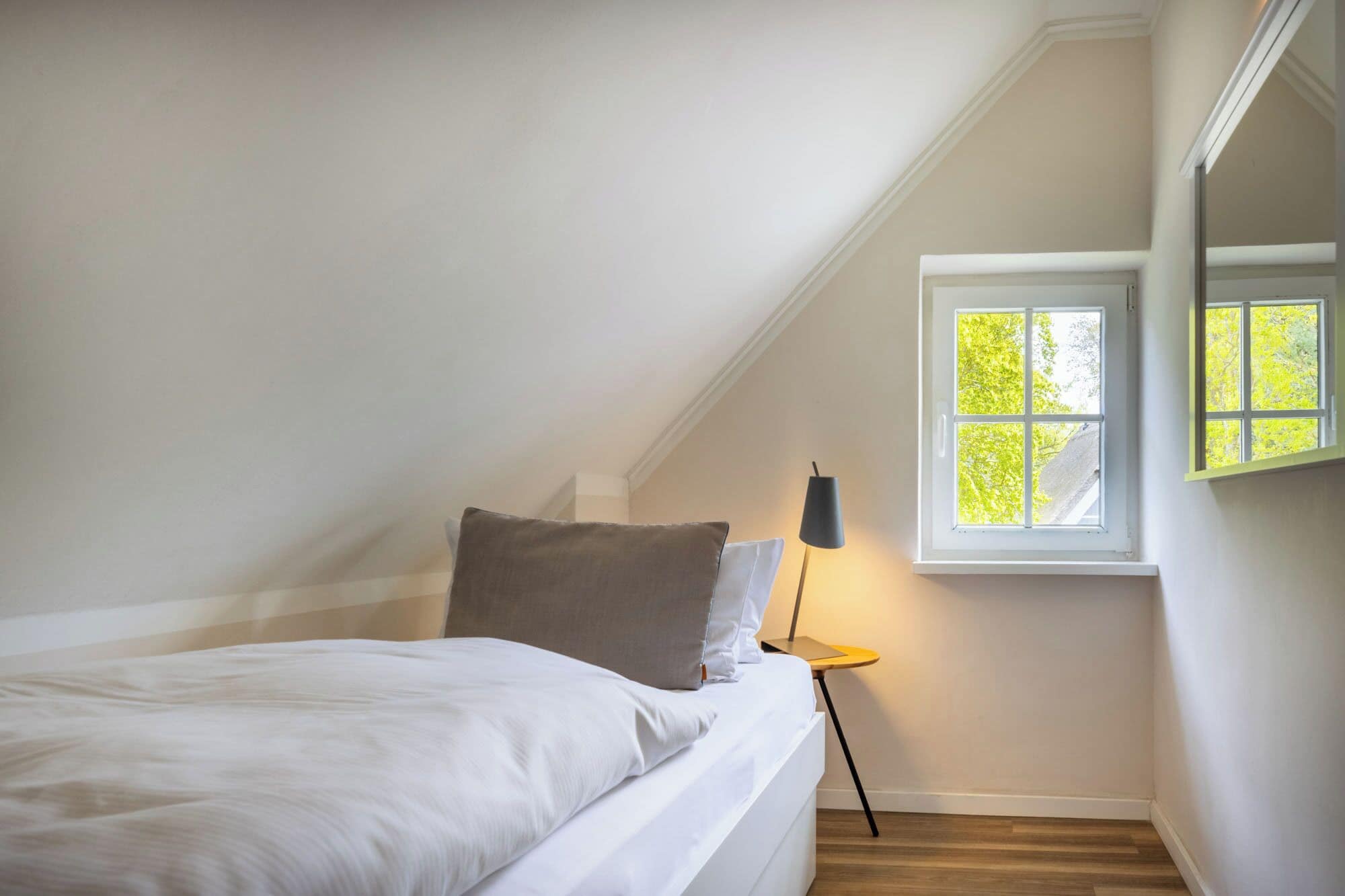 Ett sovrum med ett sluttande tak, en säng med vita sängkläder, ett sängbord med en lampa och ett fönster med utsikt över träd.