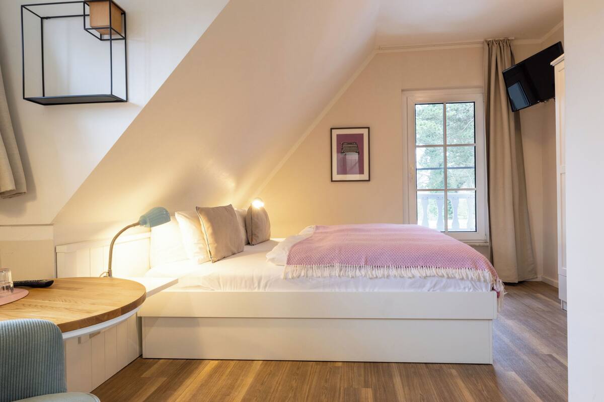 A bedroom with a bed, a desk, a lamp, a window, and a TV mounted on the wall.