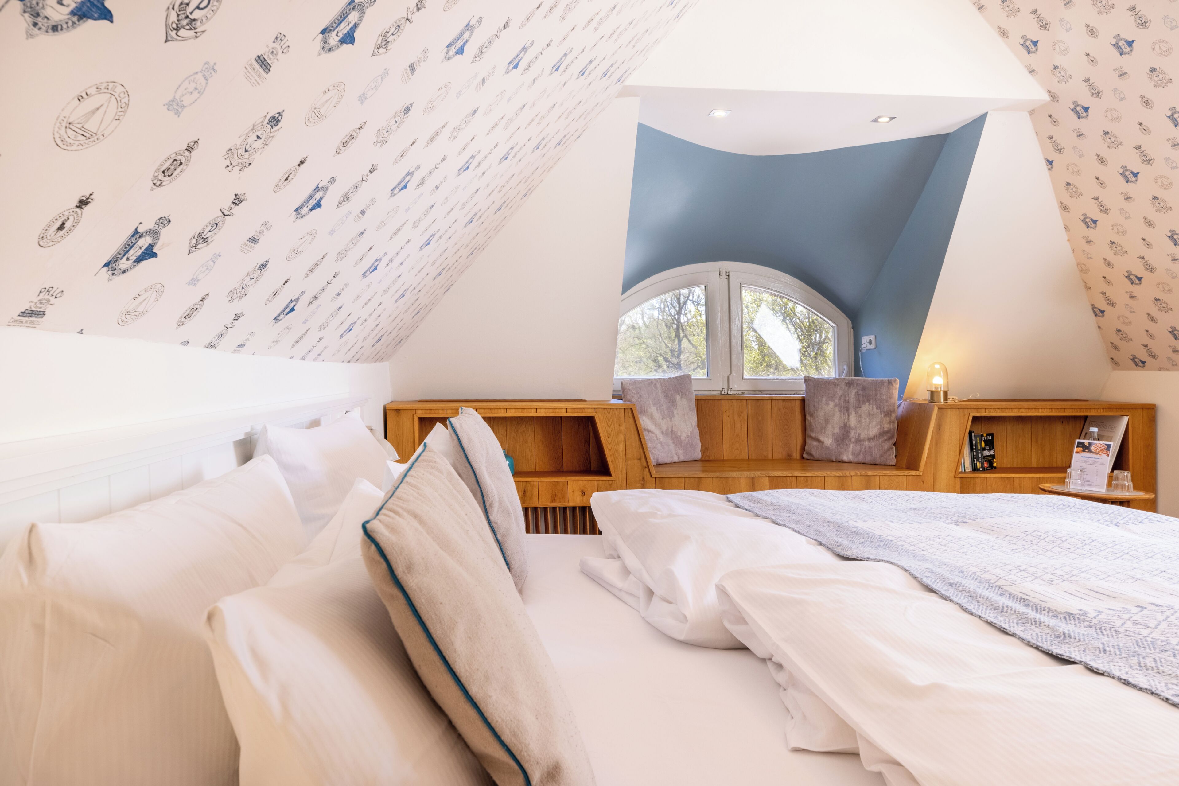 A bedroom with a sloped ceiling, a large window, wooden furniture, and patterned wallpaper.