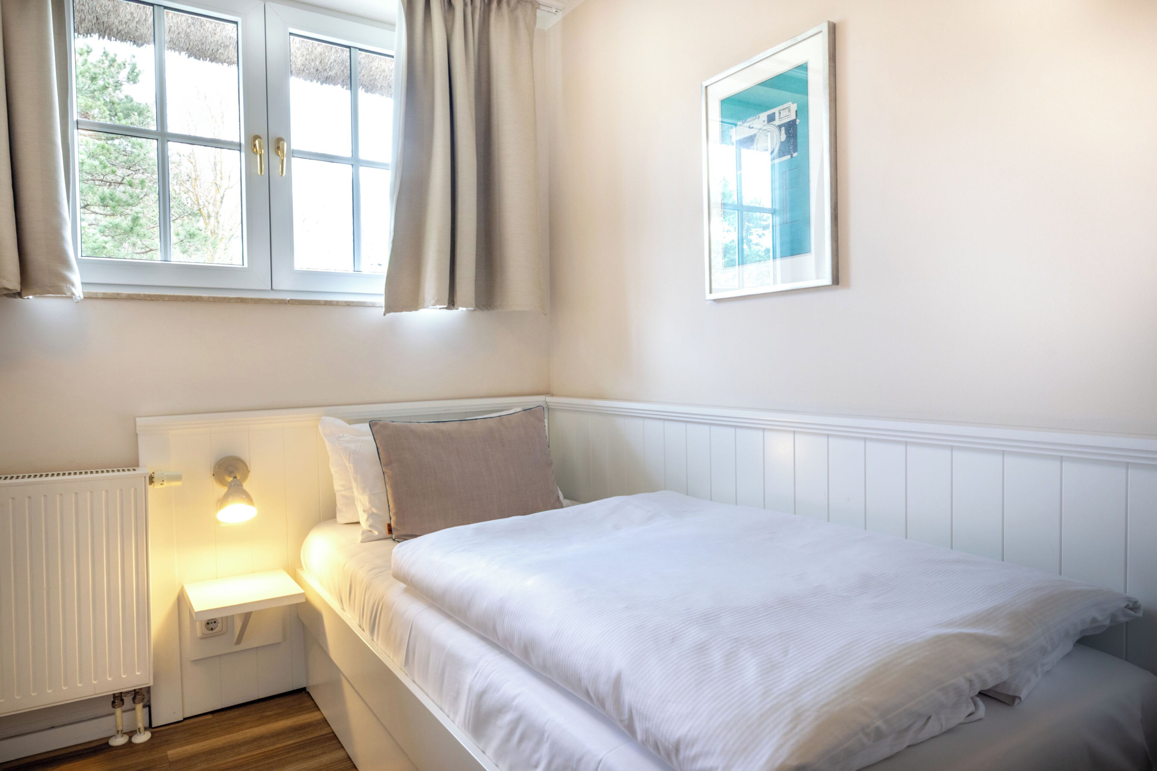 A bedroom with a bed, a nightstand, a wall-mounted lamp, a window with curtains, and a framed picture on the wall.