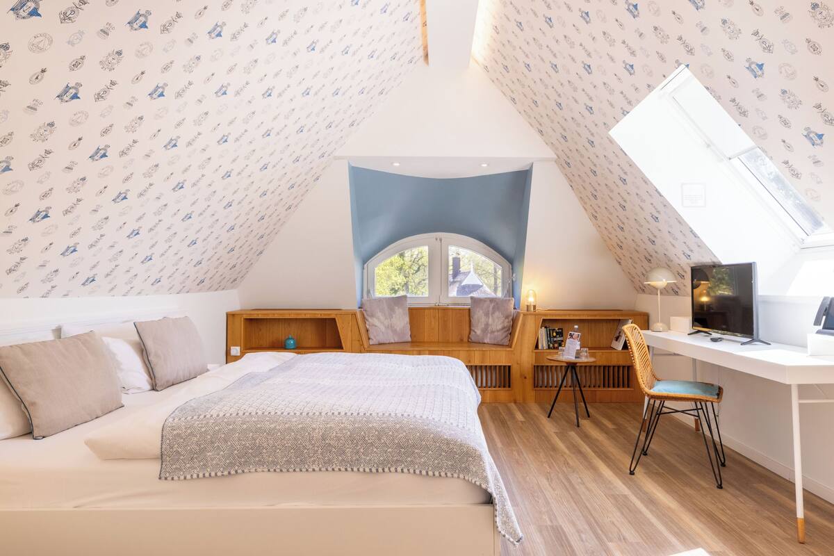 A bedroom with a bed, a desk, a chair, a TV, and a window.