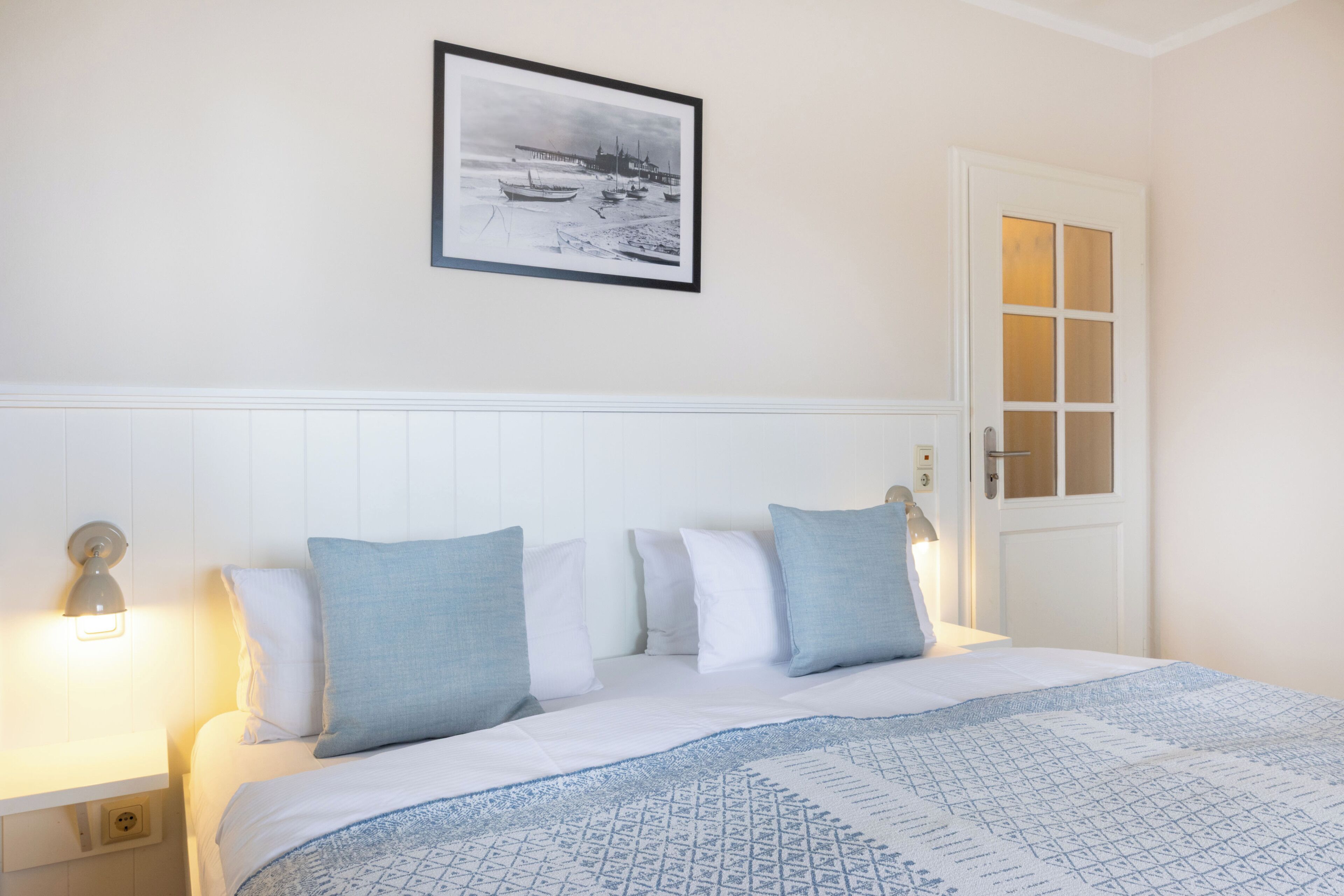 A neatly made bed with a patterned bedspread, a framed picture above it, and a white door with a glass panel.
