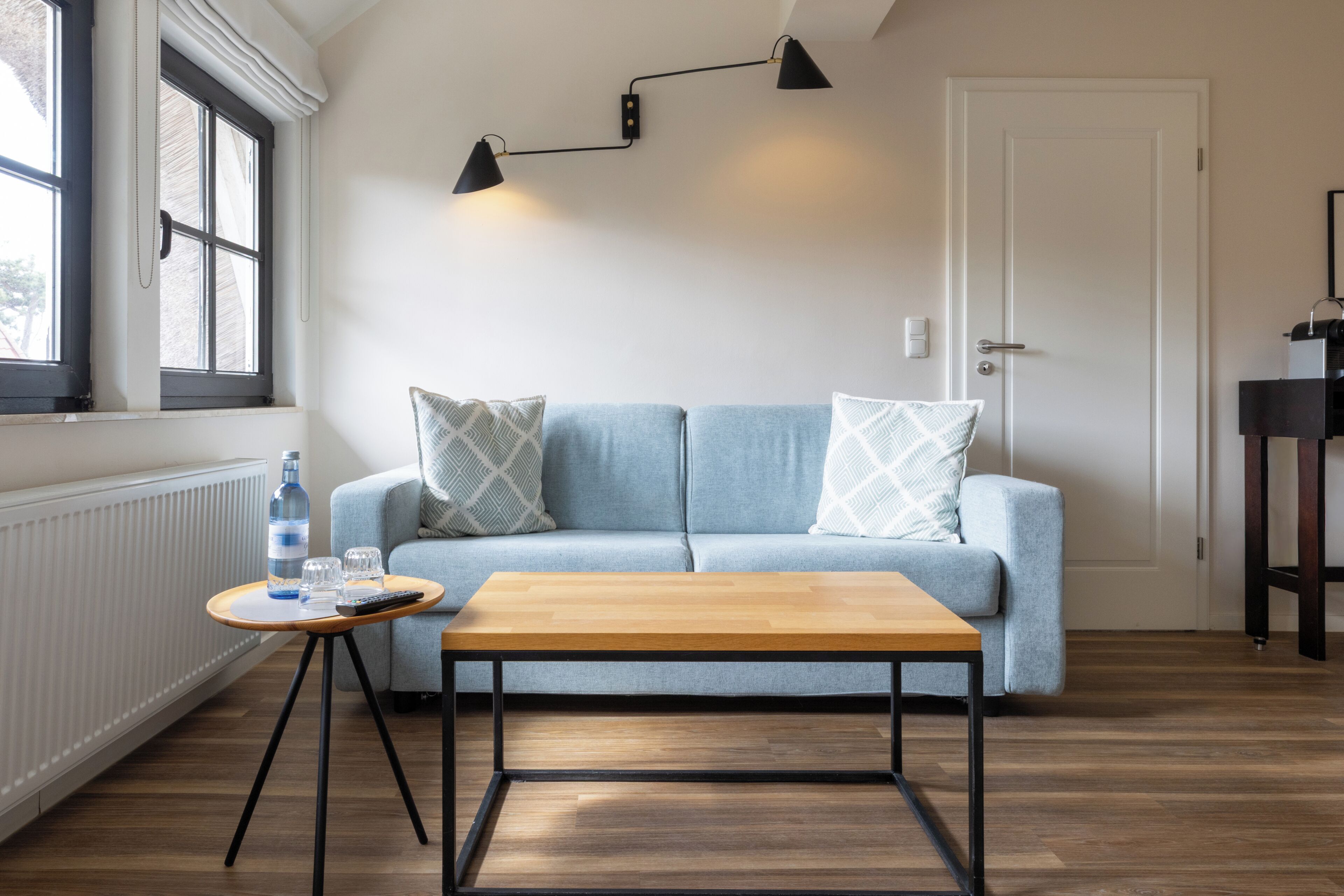 Una habitación con un sofá azul claro, una mesa de centro de madera, una pequeña mesa redonda con una botella y vasos, y una lámpara de pie negra.