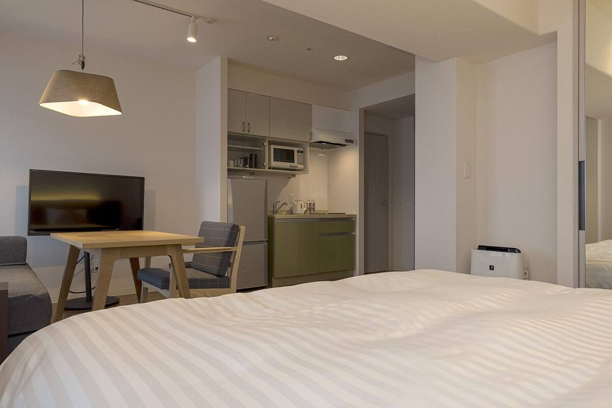 A modern hotel room with a large bed, a desk with a chair, a television, a kitchenette, and a wardrobe.