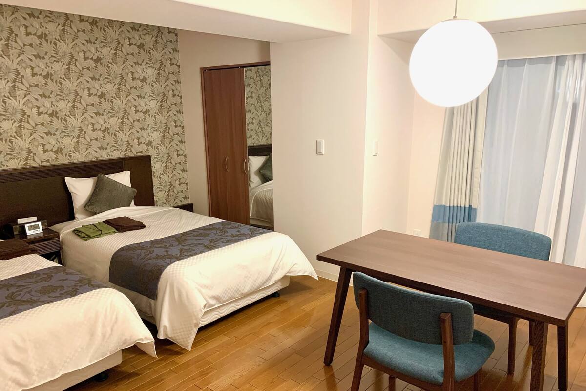 A hotel room with two beds, a wooden table, a blue chair, and a large window with curtains.