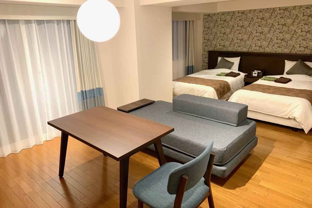A hotel room with a wooden floor, a sofa, a table, two beds with pillows and blankets, and a large window with curtains.
