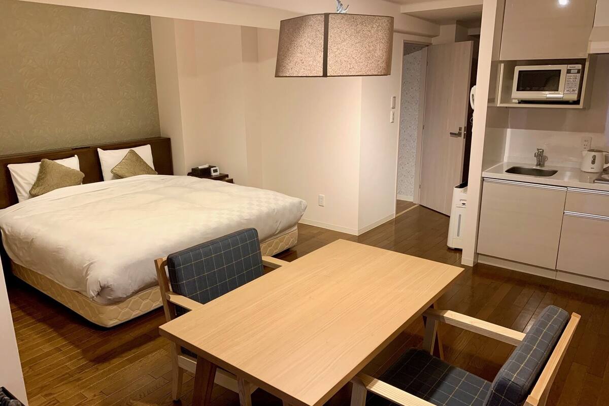 A hotel room with a bed, a dining table with chairs, a kitchen area with a microwave, and a hanging lamp.