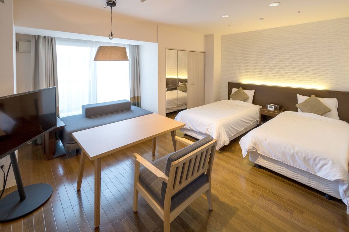 A hotel room with a wooden floor, a desk with a chair, two beds, a television, and a large window with curtains.