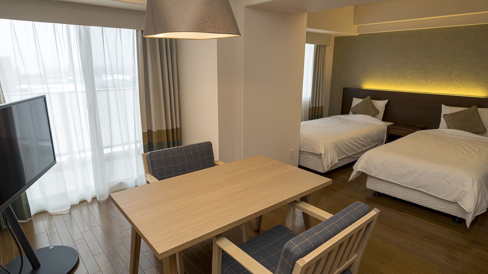 Une chambre d’hôtel avec une table en bois, deux lits, un téléviseur et une grande fenêtre avec des rideaux légers.
