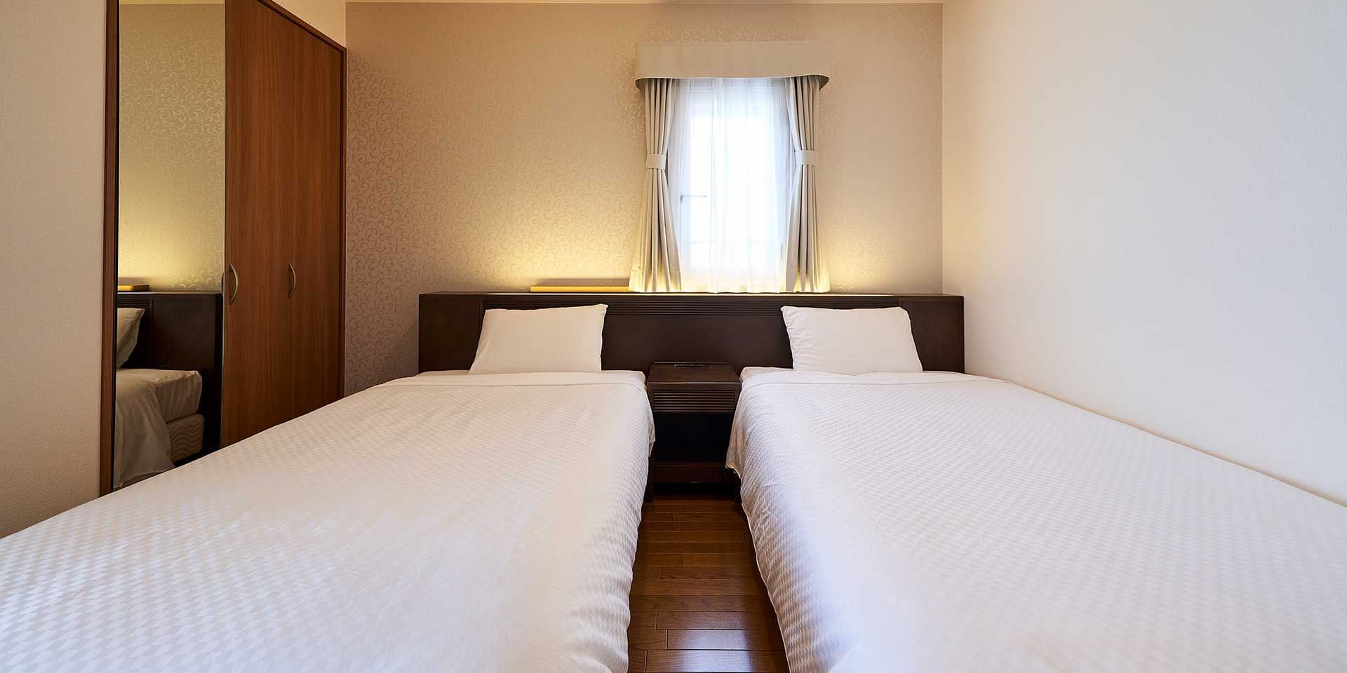 A hotel room with two single beds, a wooden wardrobe, a bedside table, and a window with curtains.