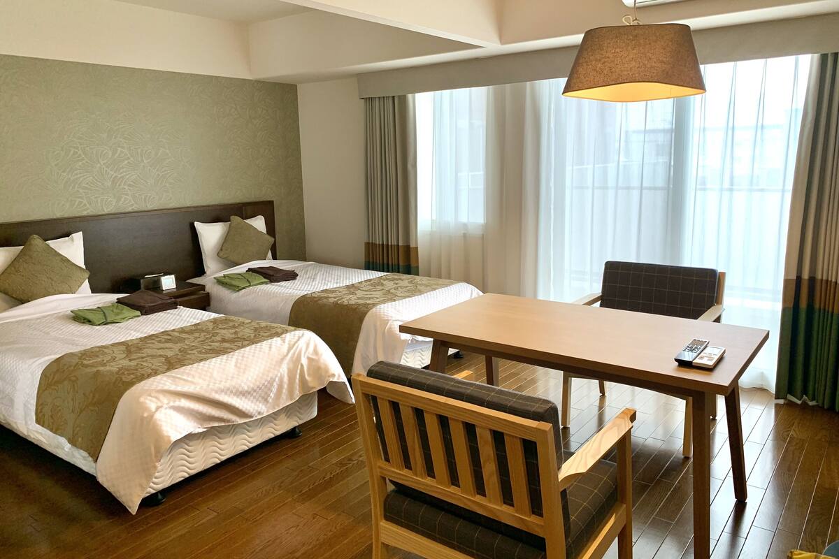 A hotel room with two beds, a wooden table, a chair, and a hanging lamp.