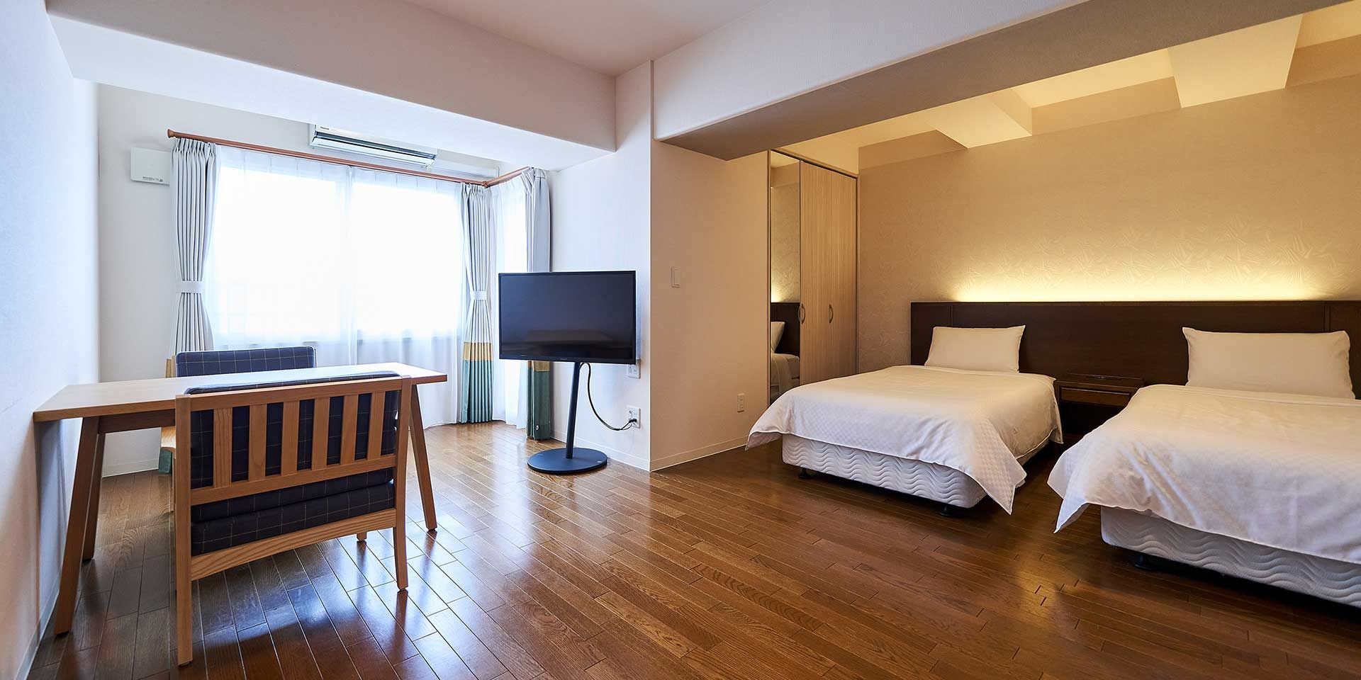 Een hotelkamer met een houten vloer, een bureau met stoel, een flatscreen-tv, twee bedden met witte lakens en een groot raam met gordijnen.