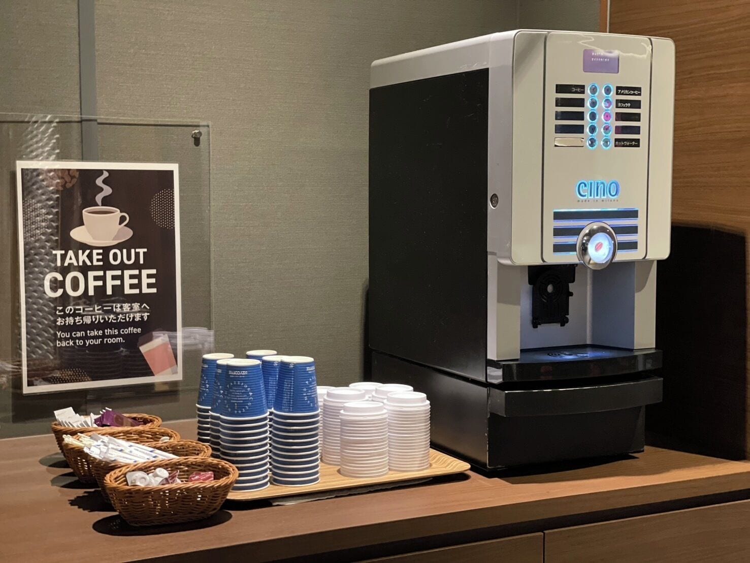 A coffee machine with a sign that says "TAKE OUT COFFEE" and stacks of disposable cups and plates.