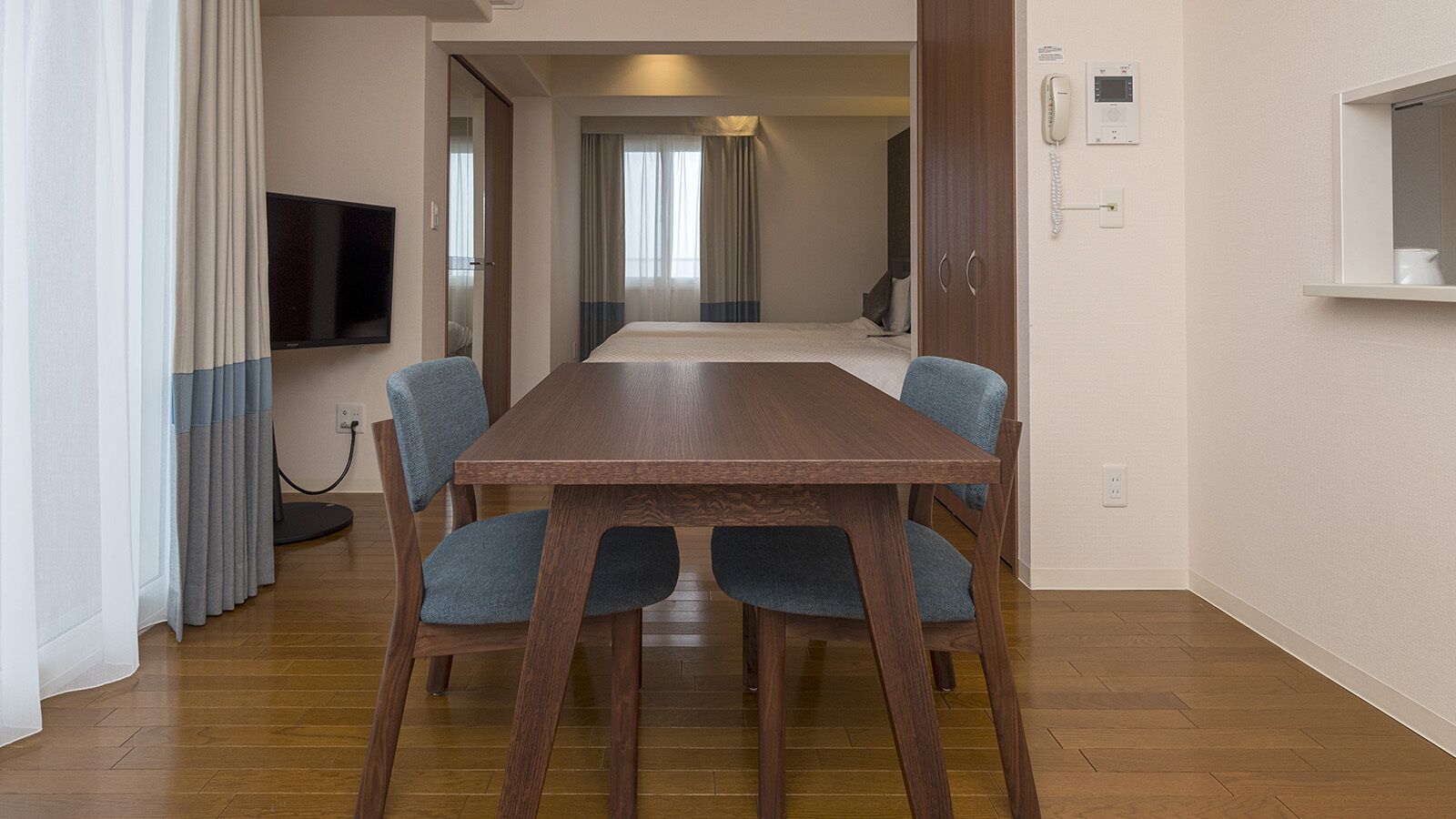 Habitación de hotel con una mesa de comedor de madera, cuatro sillas tapizadas en azul, un televisor de pantalla plana montado en la pared, una mesita de noche con lámpara y una ventana con cortinas.