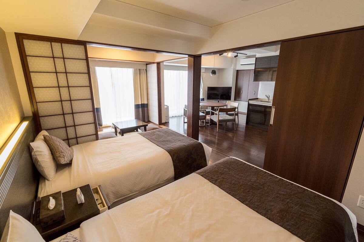 A hotel room with two beds, a desk, a chair, a TV, and a kitchenette.