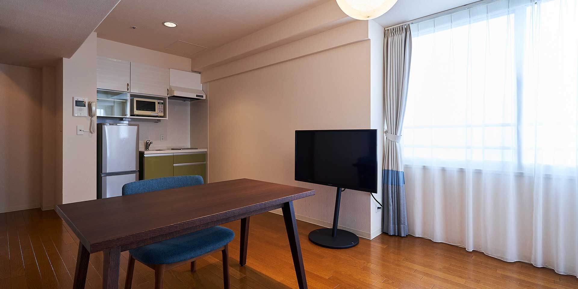 Un espacio habitable funcional con mesa, sillas, televisor de pantalla plana, kitchenette y un amplio ventanal con cortinas.