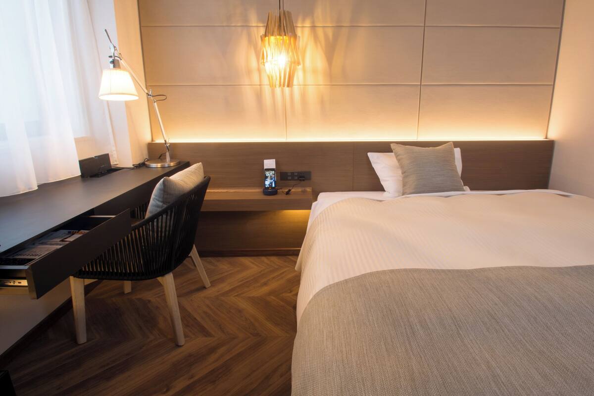 A hotel room with a bed, a desk, a chair, a nightstand, and a wall-mounted lamp.