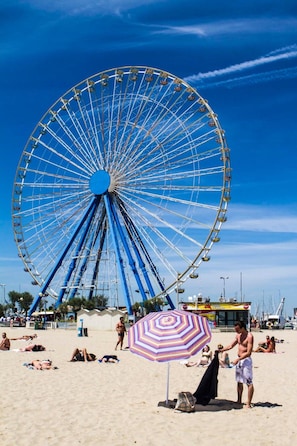 A beach scene with a large Ferris wheel, people sunbathing, and a striped umbrella. - Hotel Nova Dhely (Rimini)