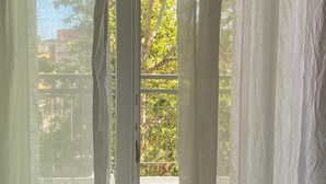 A window with sheer white curtains, a view of a tree-lined balcony, and a brick wall. - Hotel Nova Dhely (Rimini)