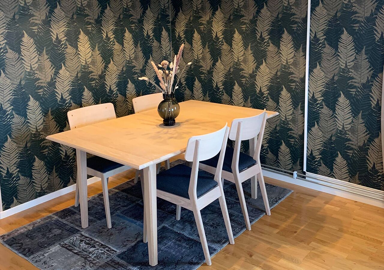 Un comedor con mesa de madera y sillas blancas, un florero con plantas secas y un fondo de papel tapiz estampado.