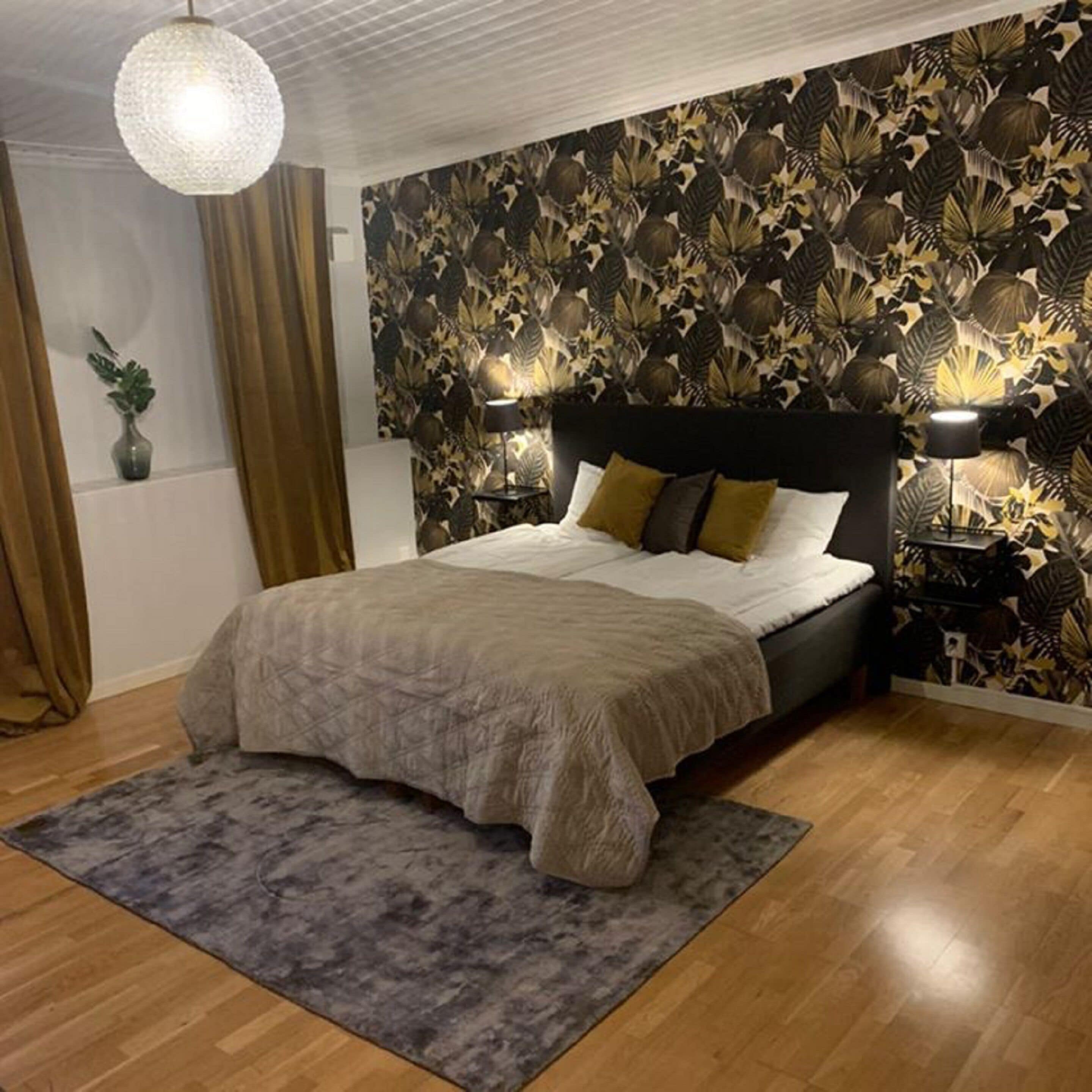 Un dormitorio con una cama grande, un tapete gris, suelo de madera, papel tapiz estampado y dos lámparas de pared.