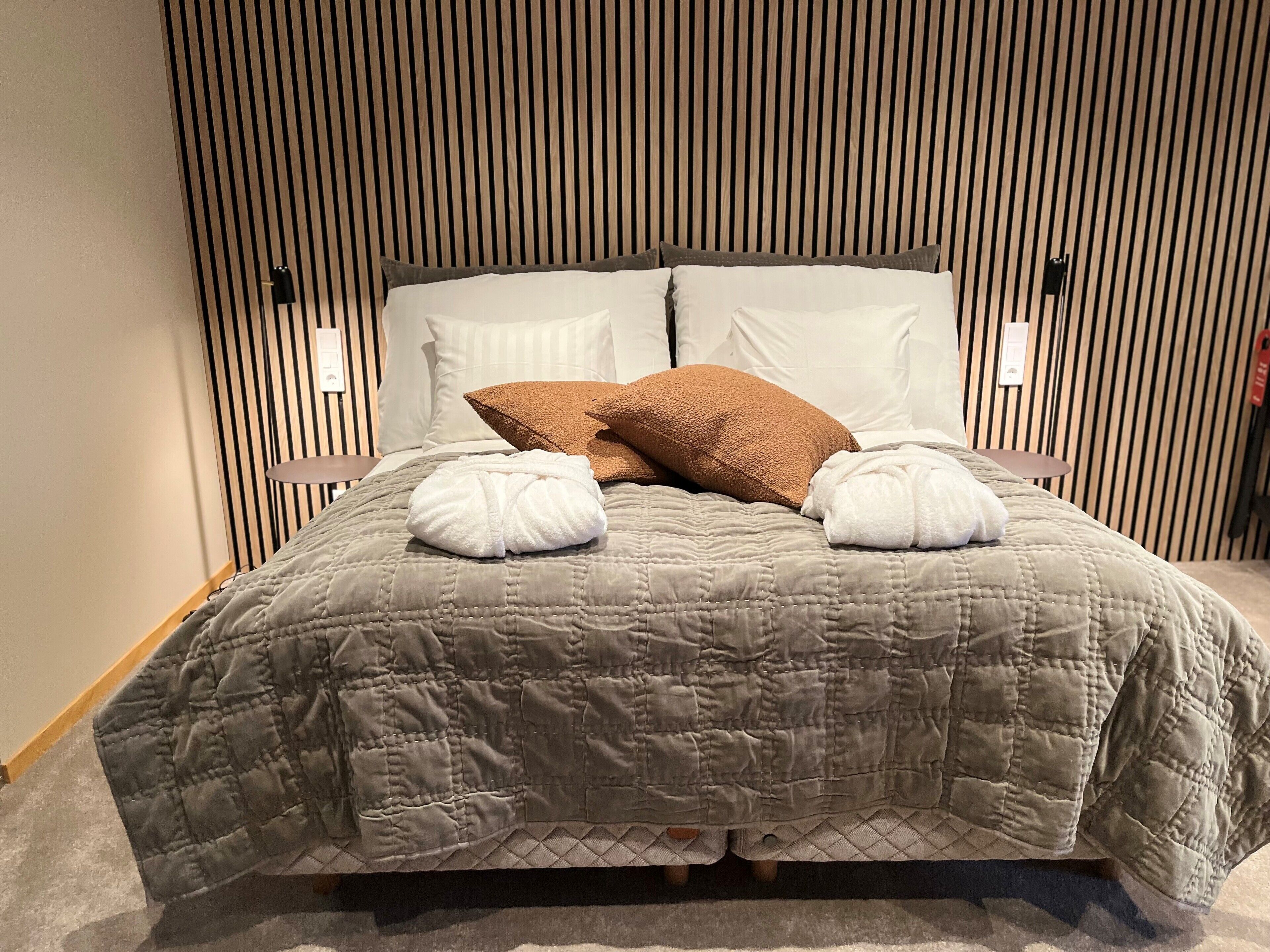 A neatly made bed with a grey quilt, two white towels, and a brown pillow.