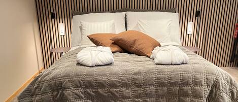 A neatly made bed with a grey quilt, two white towels, and a brown pillow.