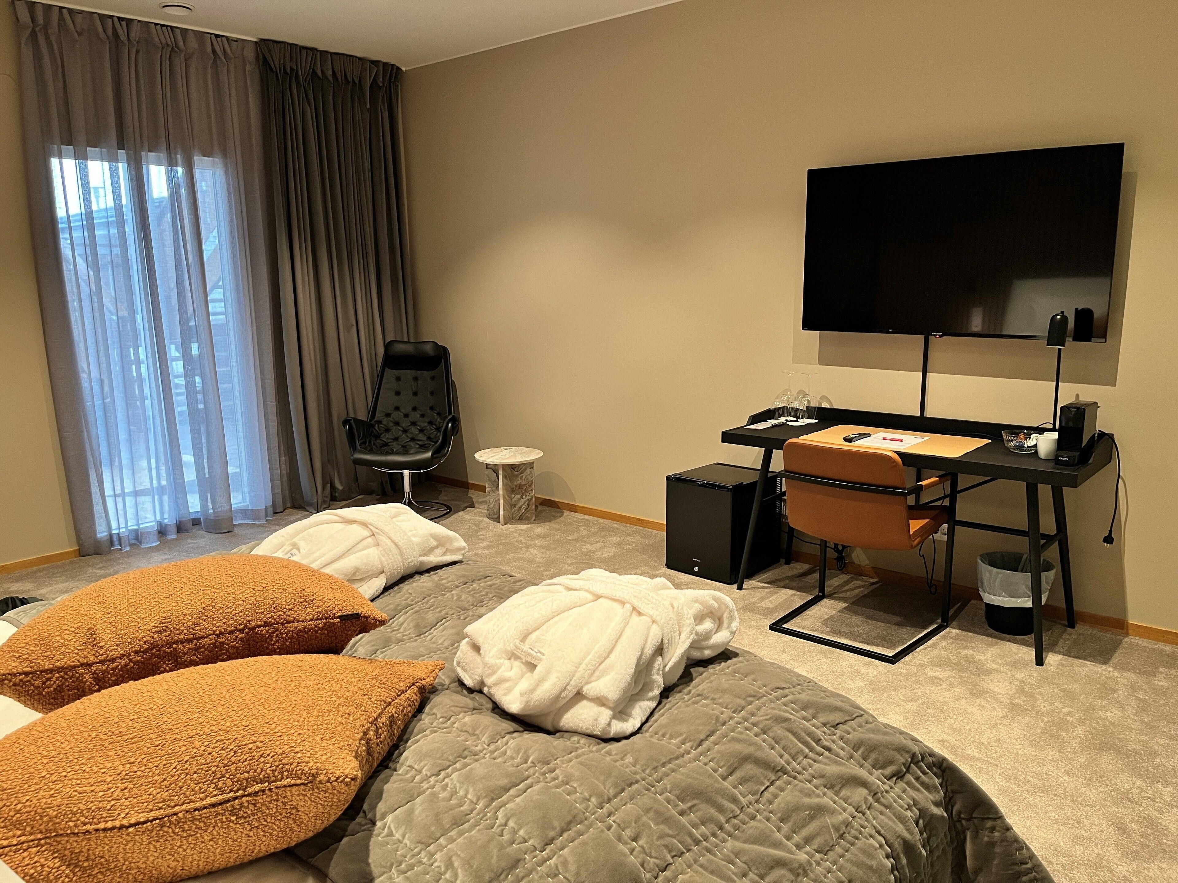 A hotel room with a bed, a desk with a computer, a chair, a TV, and a window with curtains.