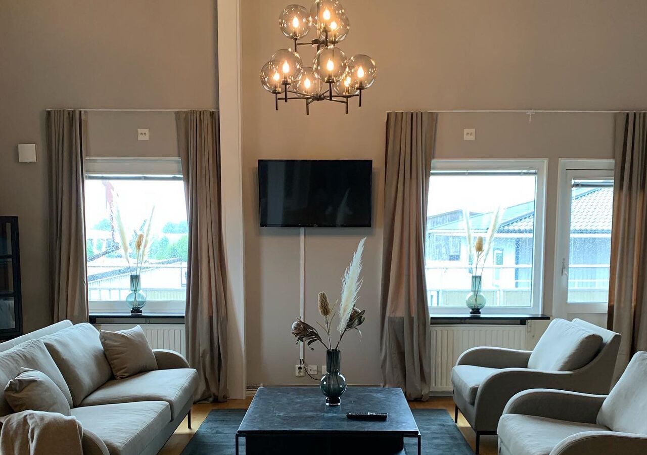 Un salon comprenant un canapé, des fauteuils, une table basse avec un vase, un téléviseur à écran plat et des fenêtres avec des rideaux.