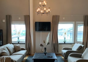 A living room with a sofa, armchairs, a coffee table with a vase, a flat-screen TV, and windows with curtains. - Hotell Ronja (Vimmerby)