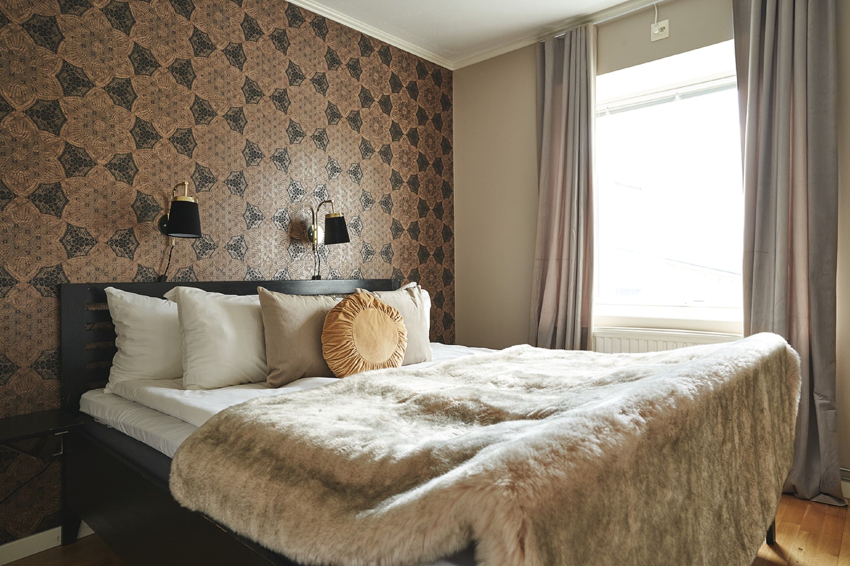 Une chambre à coucher avec un papier peint à motifs, un lit recouvert d’une couverture moelleuse, deux lampes de chevet et une fenêtre avec des rideaux.