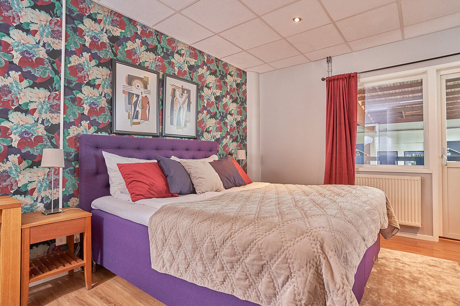 Un dormitorio con cabecera morada, papel tapiz floral, cama con varios almohadones, una mesita de noche y una ventana con cortina roja.