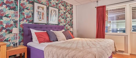 Quarto com cabeceira roxa, papel de parede floral, cama com várias almofadas, mesa de cabeceira e janela com cortina vermelha.