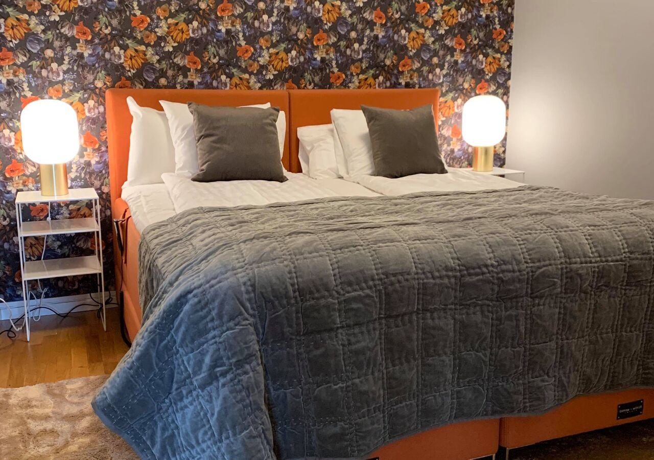 Una cama con colcha gris, dos lámparas de noche y papel tapiz con motivos florales.