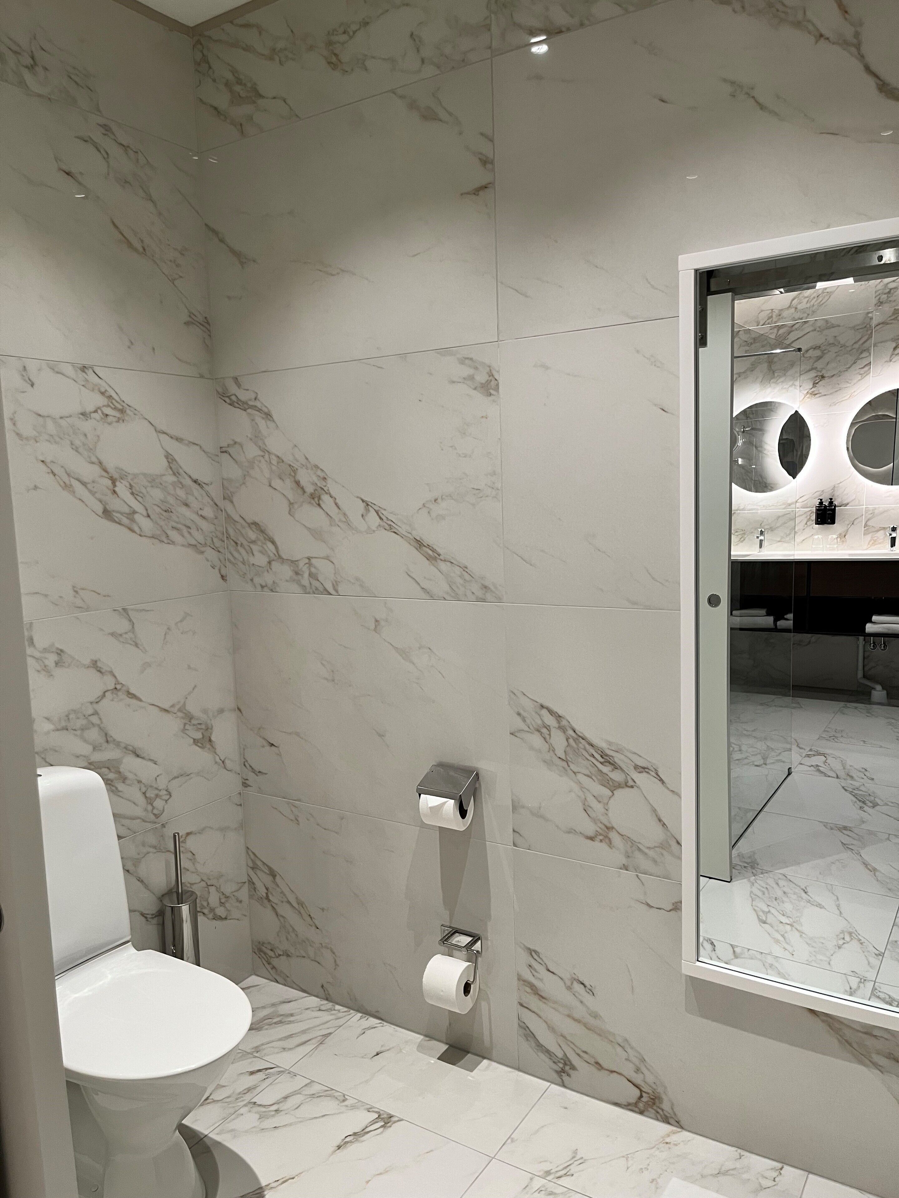 Een moderne badkamer met marmeren wanden en vloer, een toilet, een papieren handdoekdispenser en een spiegel met ronde verlichting.