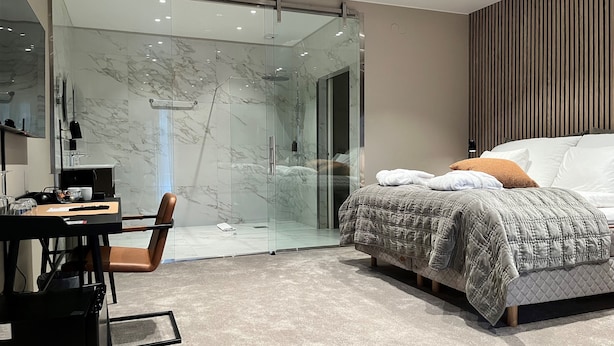 A modern bedroom with a glass shower, a bed with a quilted duvet, and a desk with a chair. - Hotell Ronja (Vimmerby)