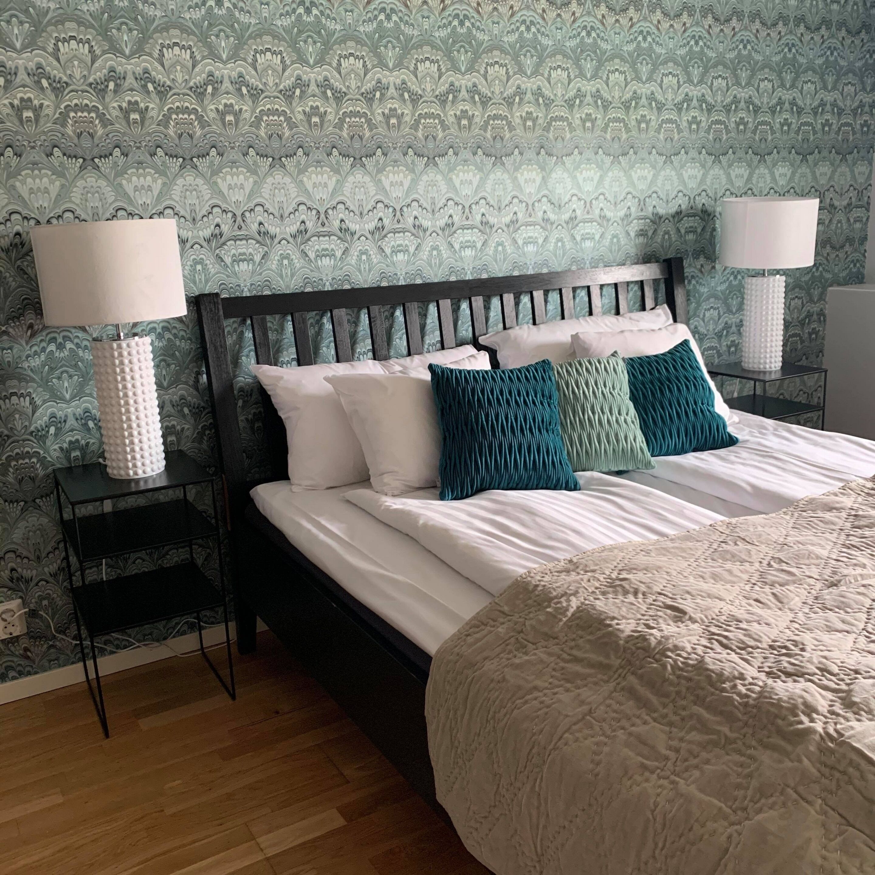 A neatly made bed with white and teal pillows, a dark wooden headboard, and a patterned wallpaper in shades of green and blue.