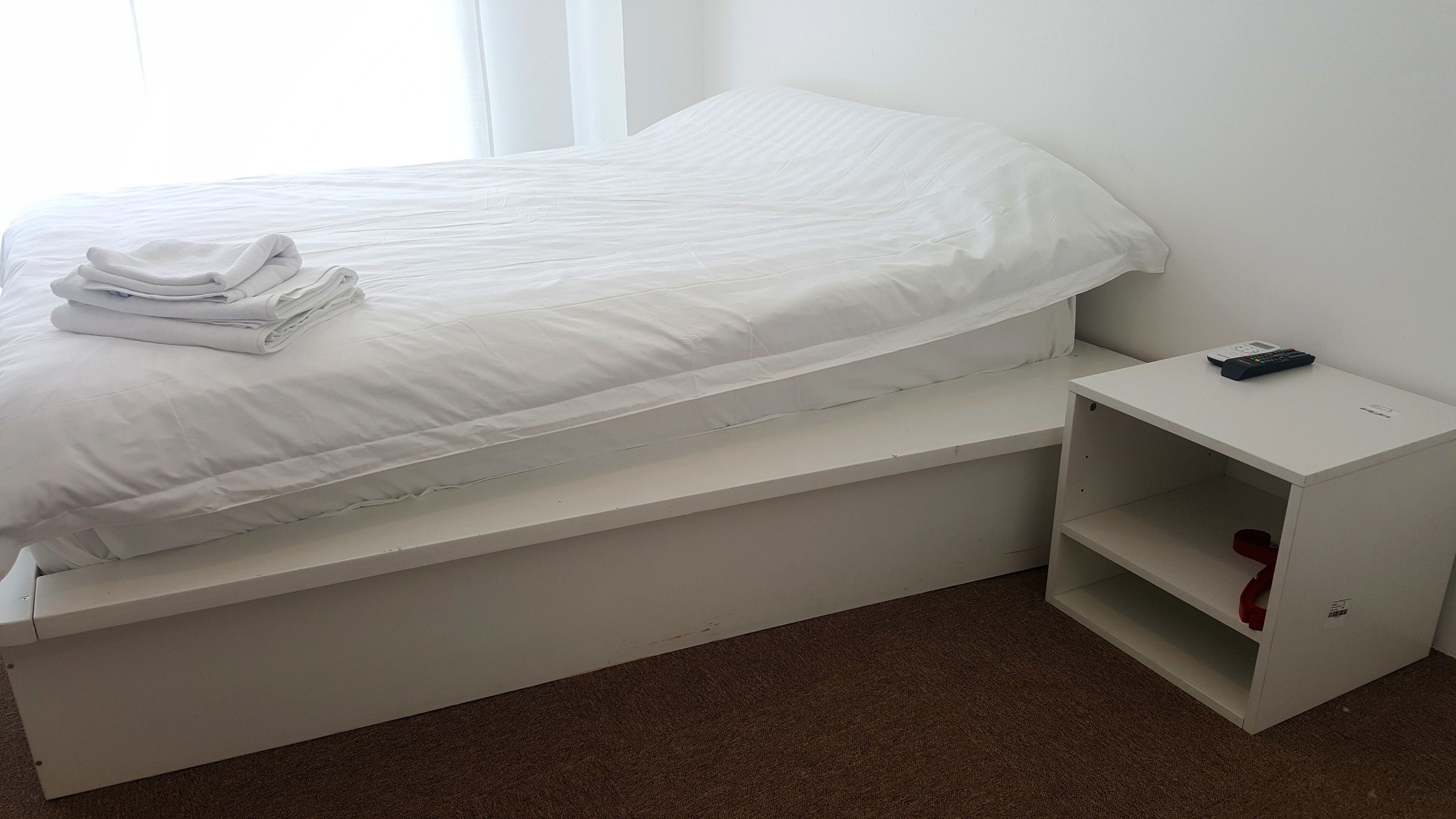 A single-bed room with a white bed, a small bedside table, and a red object on the floor.