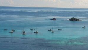Vista costera con varias embarcaciones ancladas en aguas tranquilas, una pequeña isla con vegetación en la distancia y un horizonte despejado.