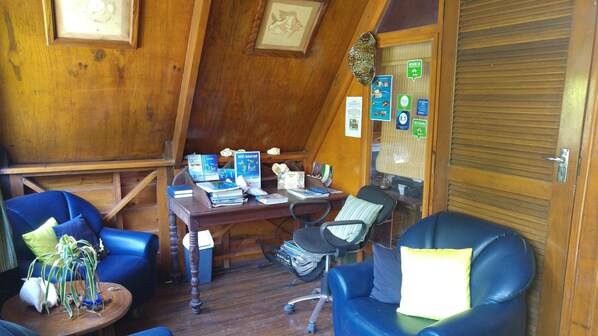 Un interior de madera con un escritorio, dos sillas azules, una pequeña mesa con una planta y una puerta cerrada con una ventana.