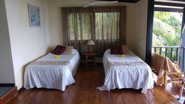 A room with two beds, a small table with a lamp, and a balcony with a chair. - Mango Lodge (Praslin Island)
