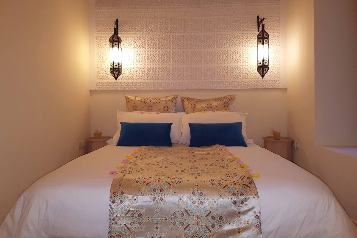 A bed with a patterned bedspread, two blue pillows, and two wall-mounted lamps.
