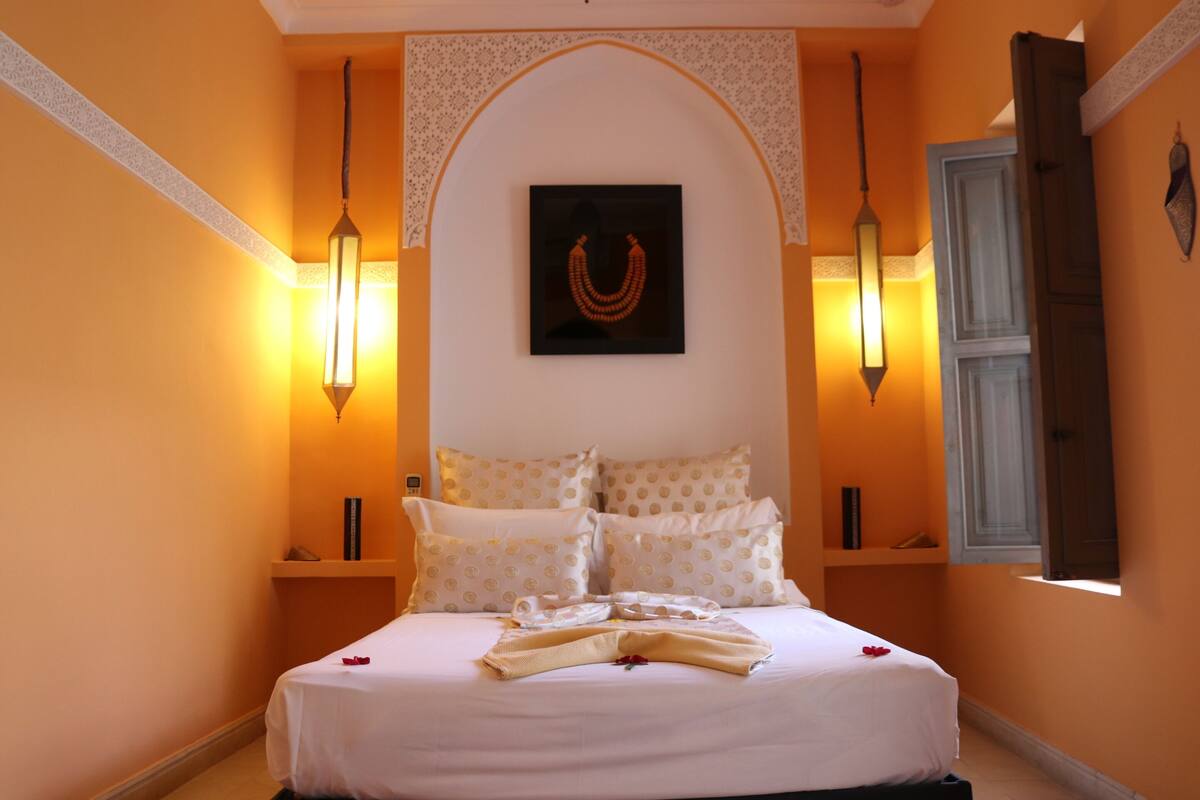 A neatly made bed with white linens and a decorative pillow arrangement, a wall-mounted picture, and two wall sconces.