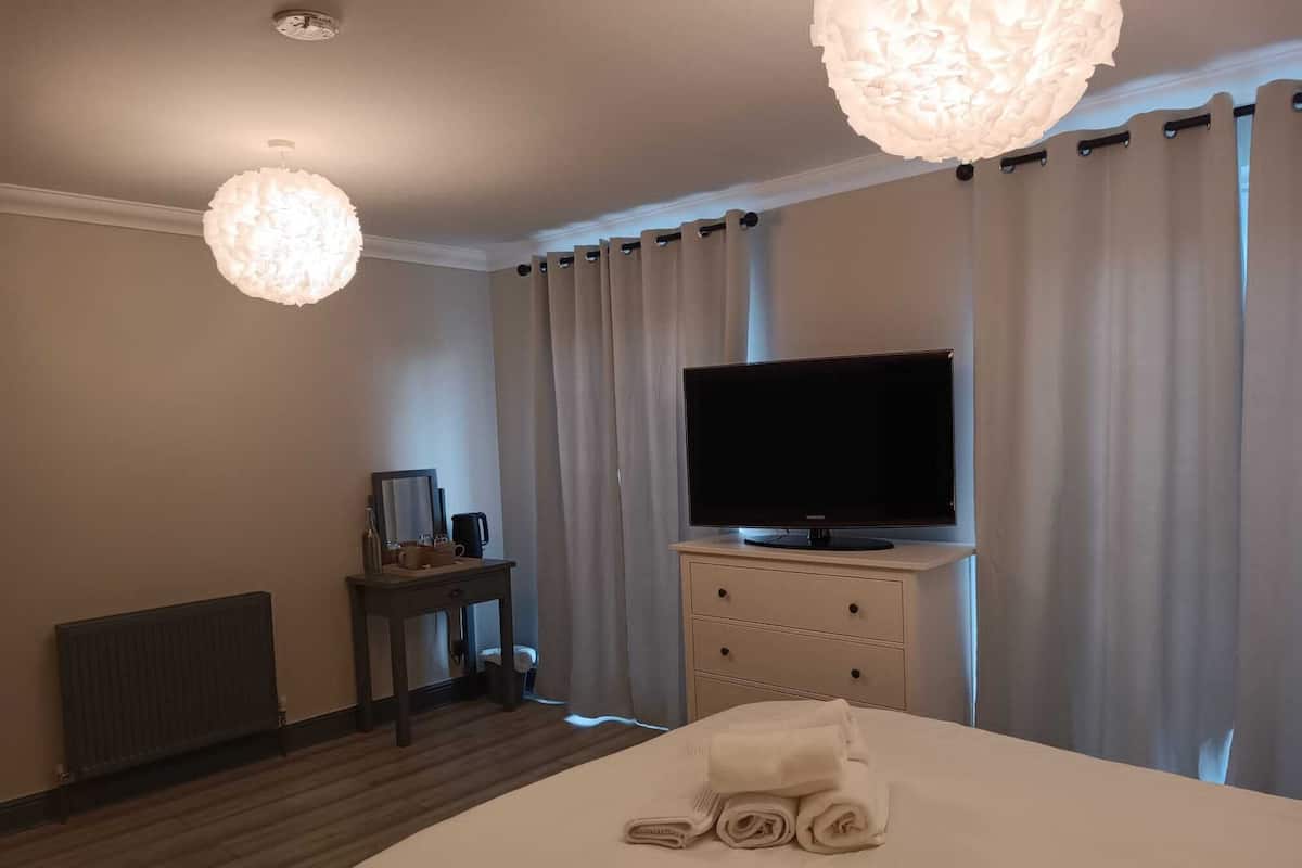 A hotel room with a bed, a television on a stand, a bedside table with a mirror, and a radiator.