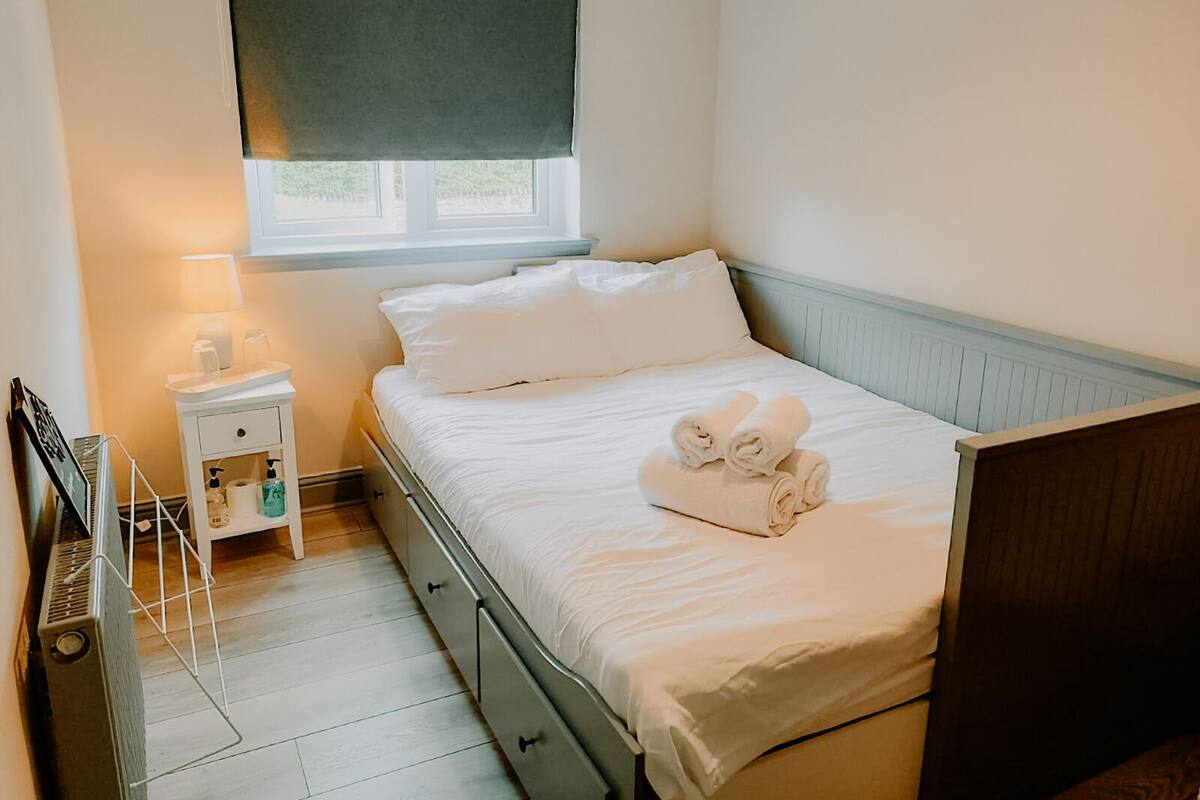 A bedroom with a bed, a bedside table with a lamp, a window with a blind, and a radiator.